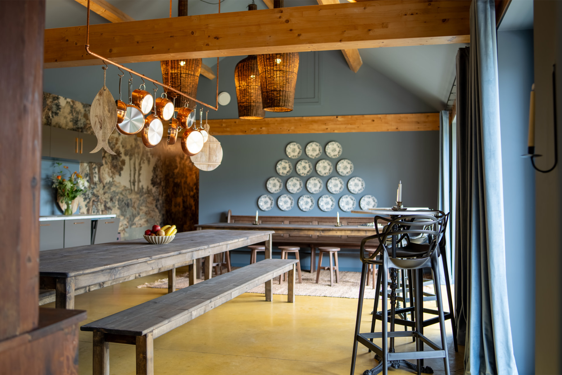 Koperen pannen aan een rek boven de lange eettafel, met rieten lampen aan de balken